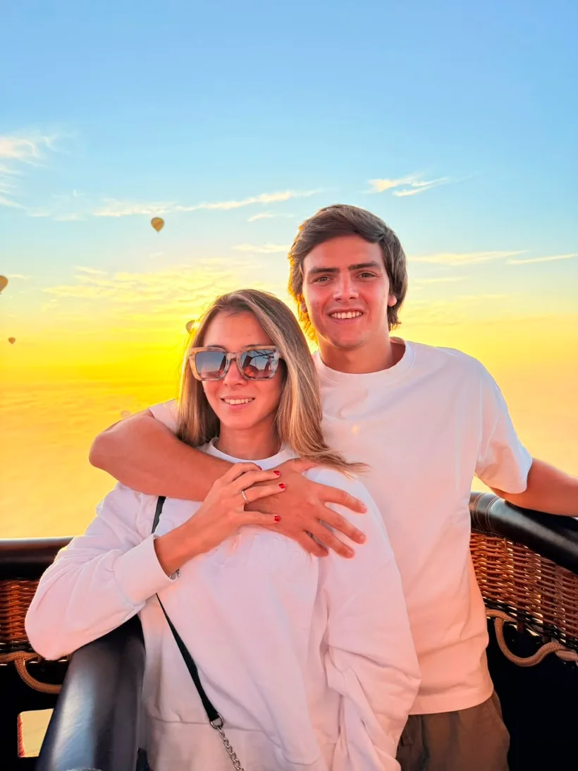 Casal abraçado no cesto do balão ao nascer do sol, com outros balões ao fundo