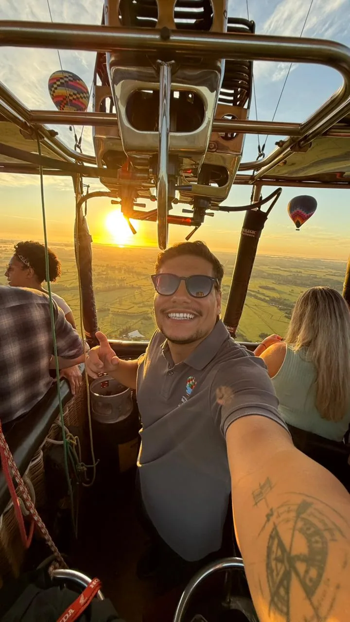 O piloto Diovane no cesto do balão, apontando para a câmera com o nascer do sol ao fundo